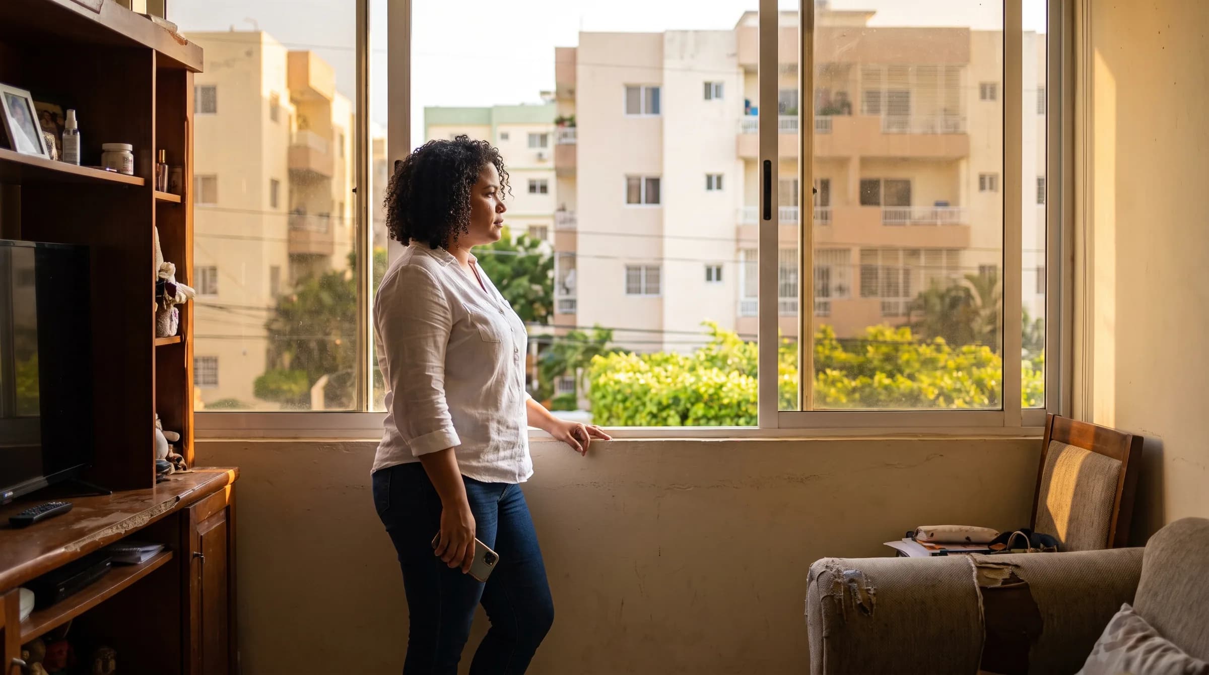Mujer dominicana joven reflexionando mientras observa por la ventana de un apartamento en La Julia, Santo Domingo, sosteniendo su teléfono a un lado