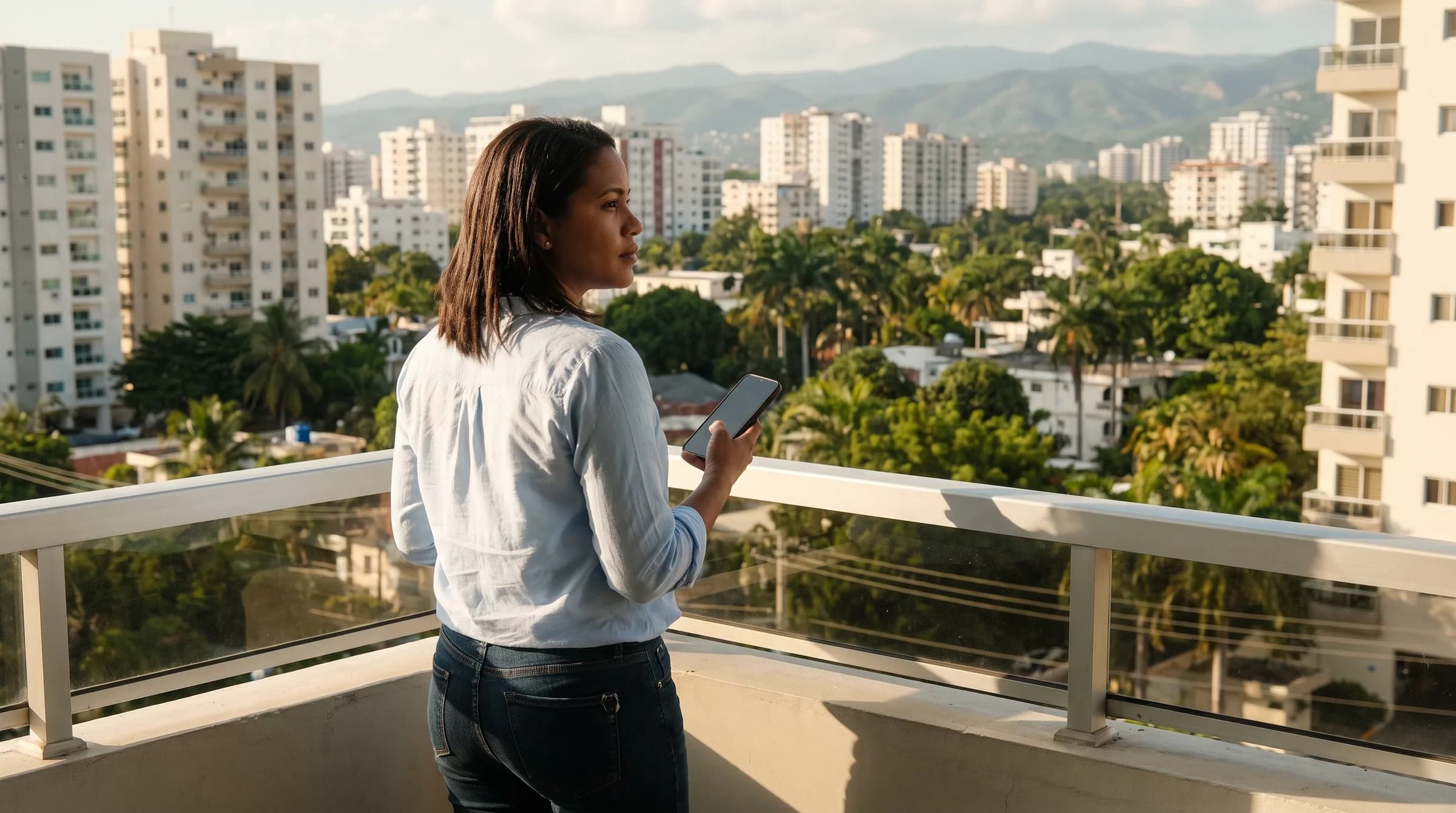 Mujer joven contemplando la vista de Santo Domingo desde el balcón de un apartamento, sosteniendo su teléfono mientras evalúa una decisión de compra inmobiliaria