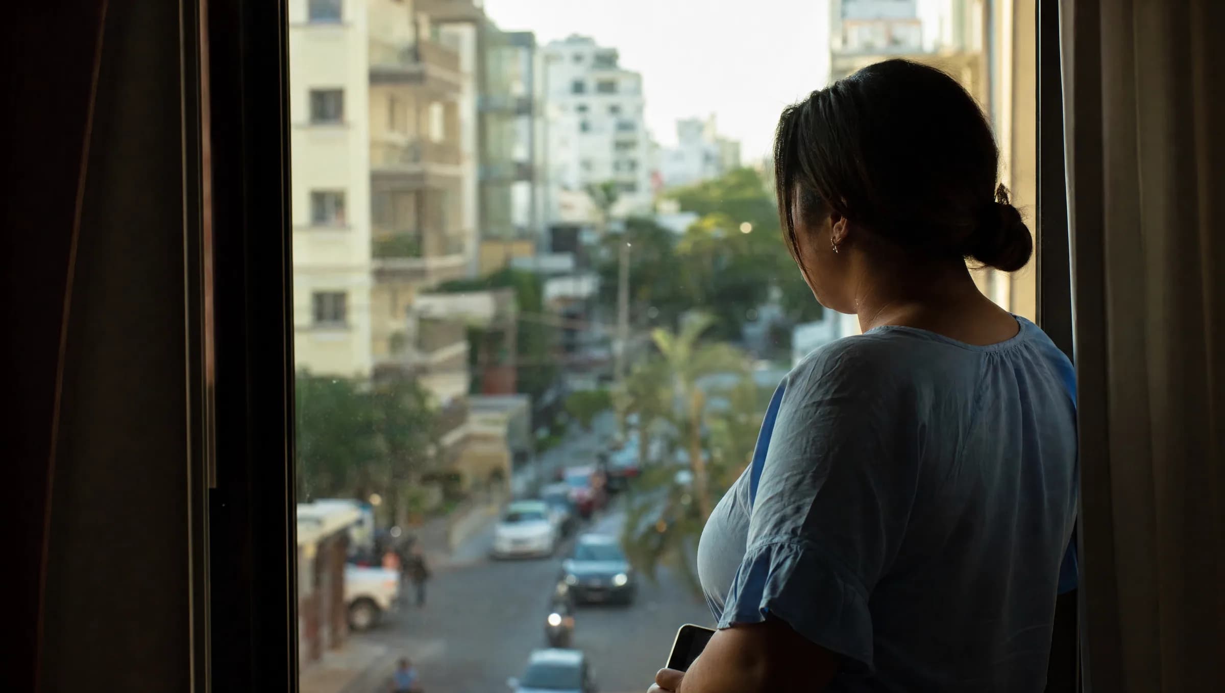 Mujer dominicana contemplando la vida urbana de Santo Domingo desde la ventana de su apartamento, reflejando sobre las realidades del costo de vida en la ciudad