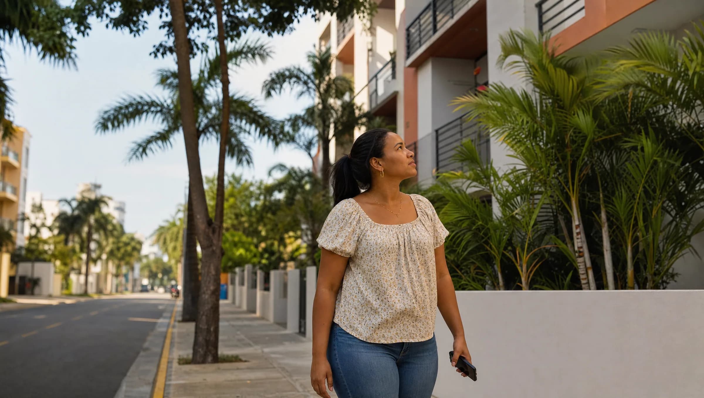 Mujer dominicana contemplando edificio de apartamentos en Santo Domingo Este mientras sostiene su teléfono, reflejando el proceso de decisión al comprar vivienda