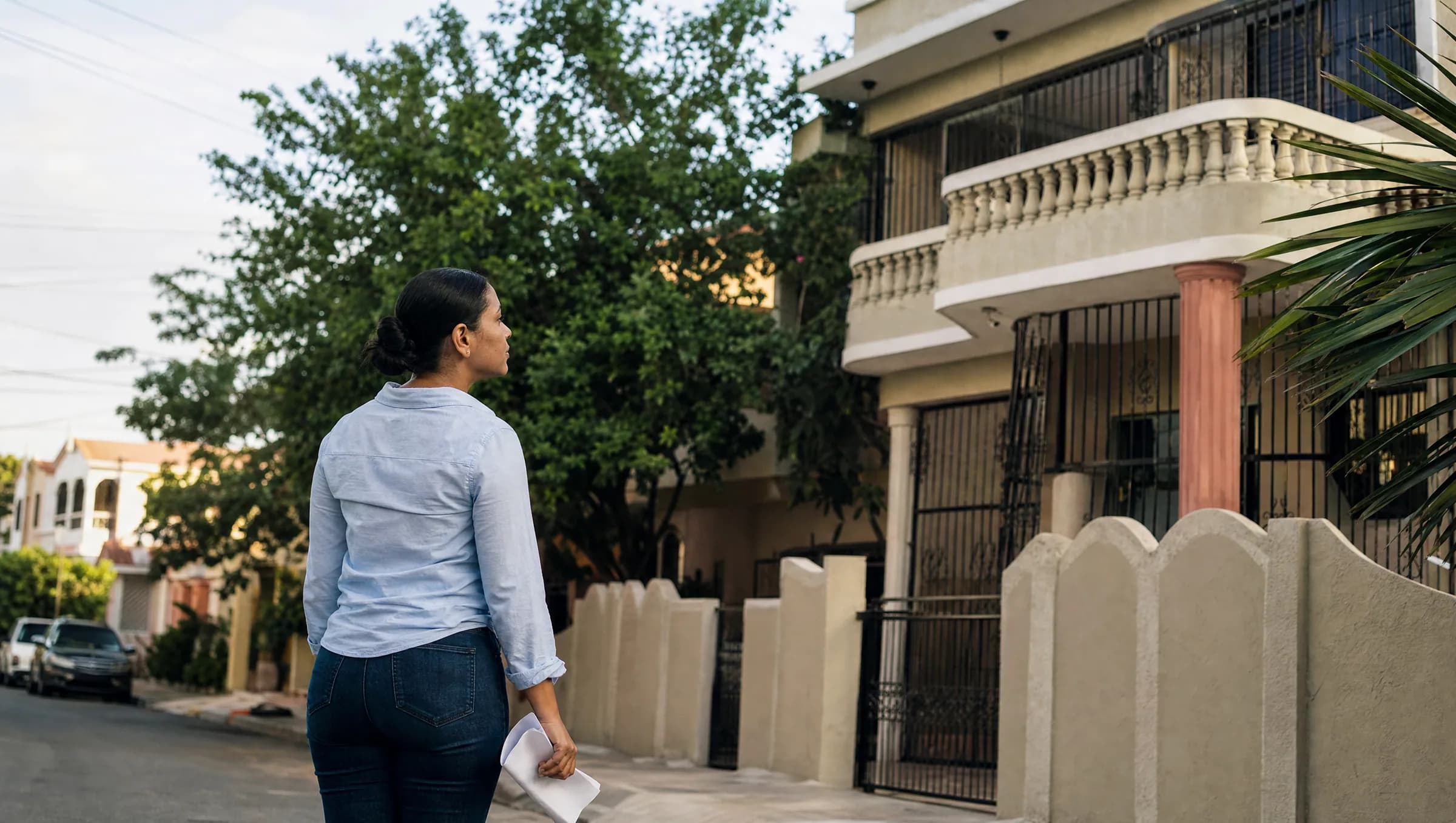 Mujer dominicana observando una propiedad residencial en Santiago mientras sostiene documentos, evaluando una posible compra de inmueble