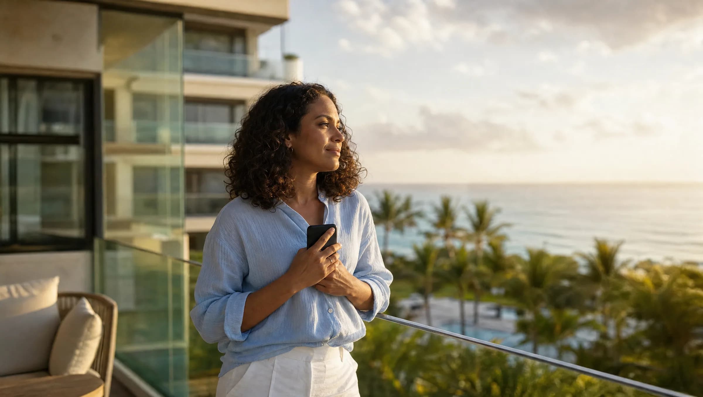 Mujer dominicana reflexionando en balcón de apartamento en Punta Cana mientras sostiene su teléfono