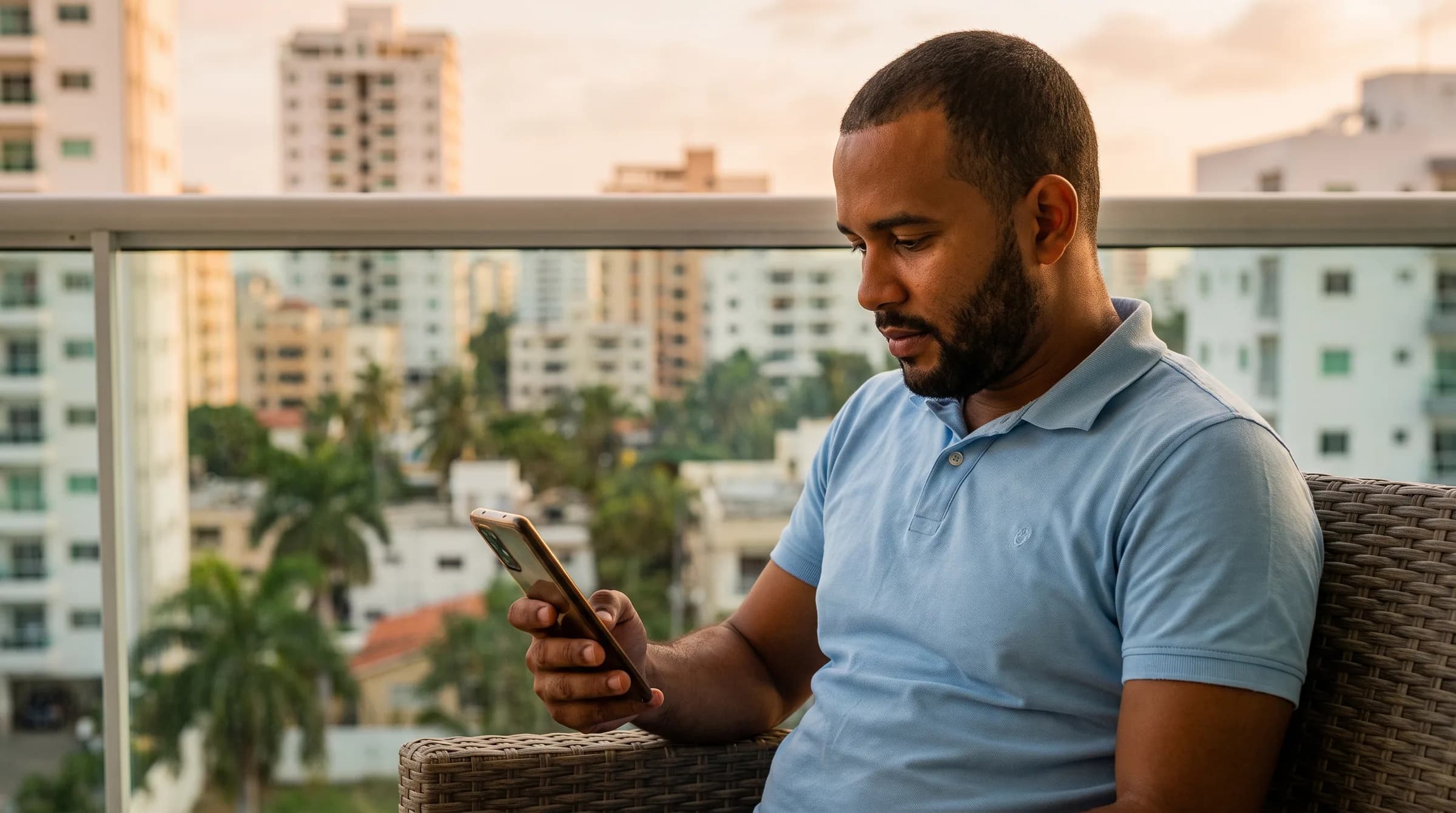 Hombre dominicano revisando información en su teléfono desde la terraza de un apartamento en Santo Domingo, con el paisaje urbano de fondo