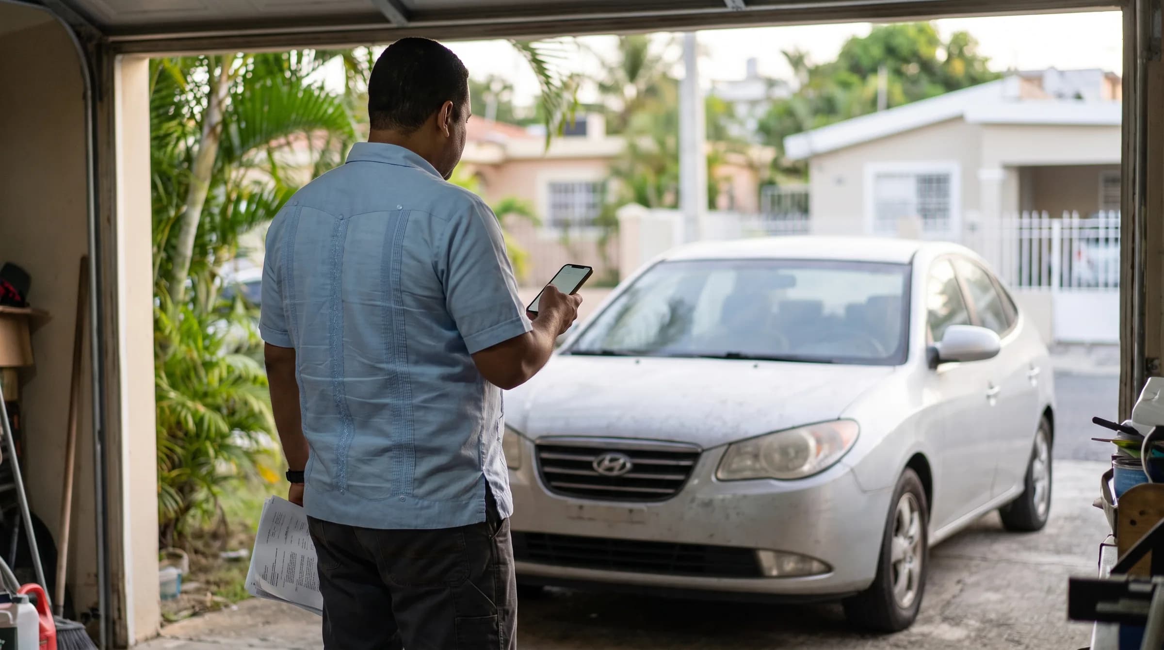 Hombre dominicano de espaldas observando su carro recién importado en el garaje de su casa, sosteniendo un teléfono mientras revisa documentos
