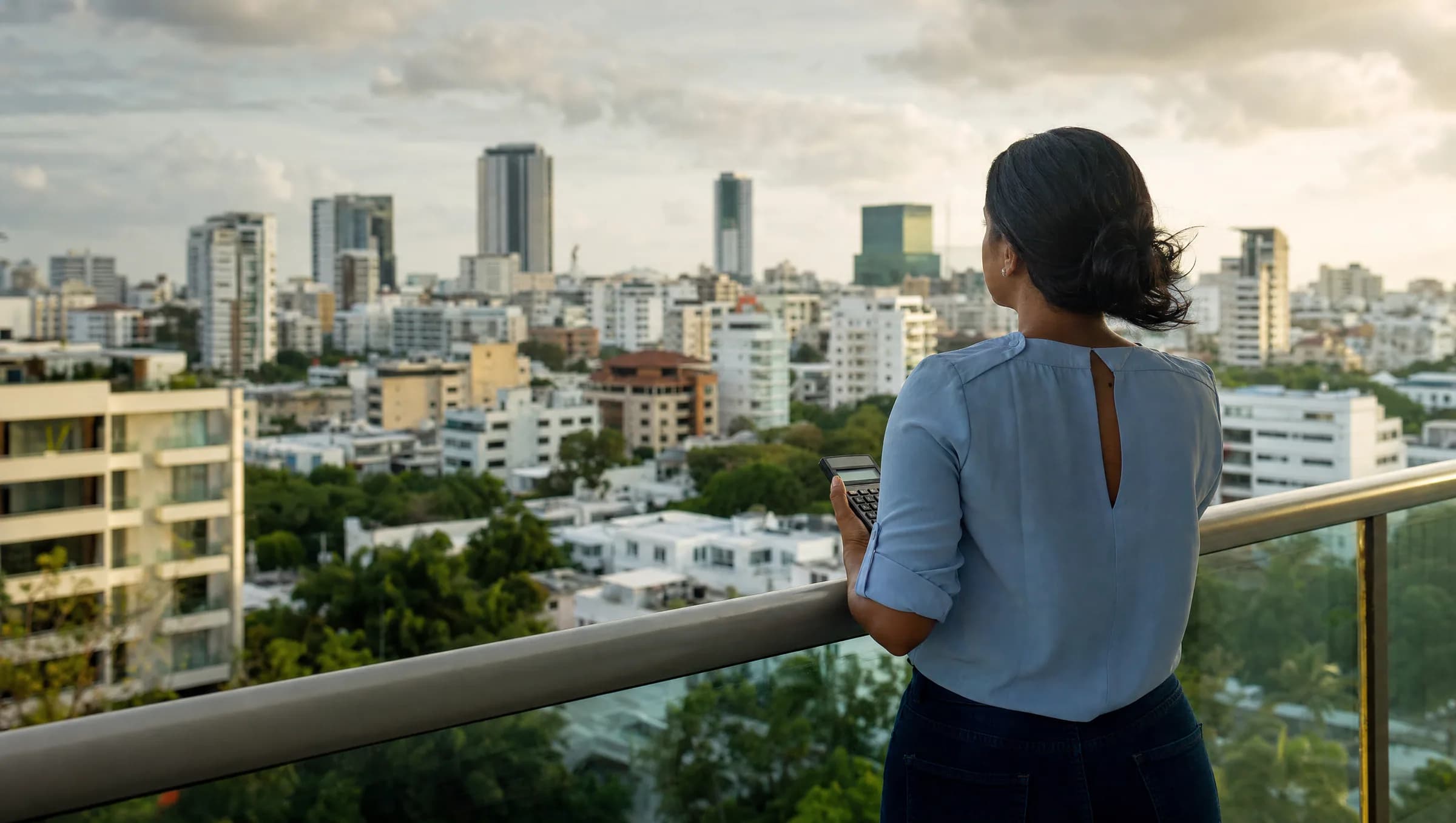 Mujer dominicana contemplando pensativa la vista de Santo Domingo desde el balcón de un apartamento, sosteniendo una calculadora mientras reflexiona sobre los costos de compra de su propiedad