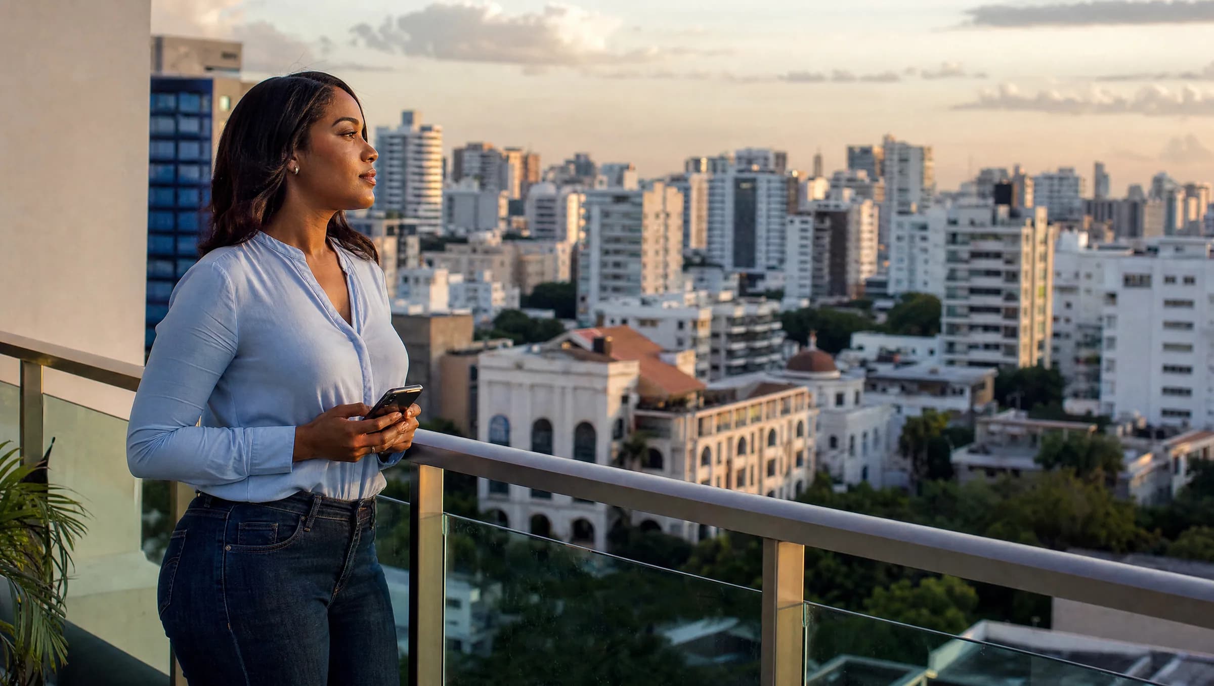 Mujer dominicana joven reflexionando en el balcón de un apartamento mientras observa el horizonte urbano de Santo Domingo