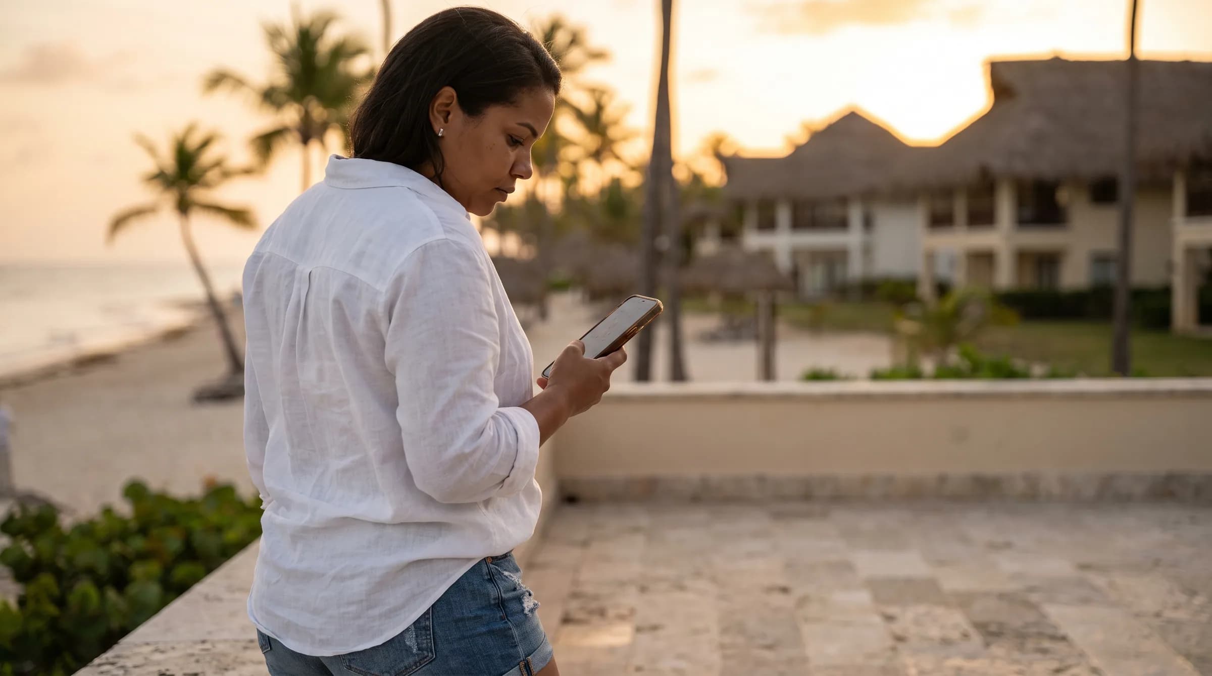Mujer dominicana reflexionando mientras revisa su teléfono en una terraza frente a la playa de Punta Cana, con edificios residenciales al fondo