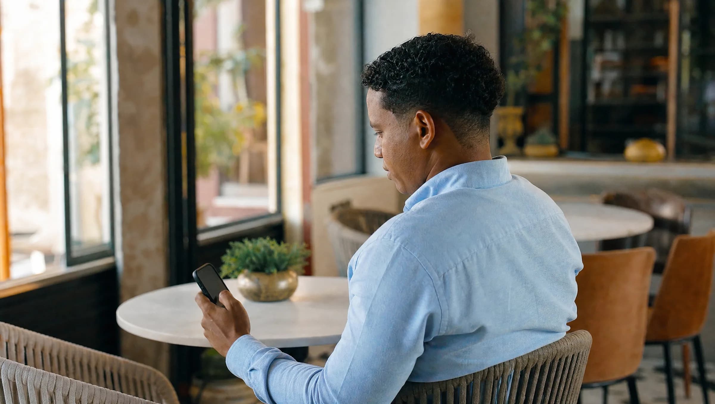 Hombre dominicano revisando mensajes en su teléfono con expresión preocupada mientras busca agente inmobiliario en una cafetería de Santo Domingo
