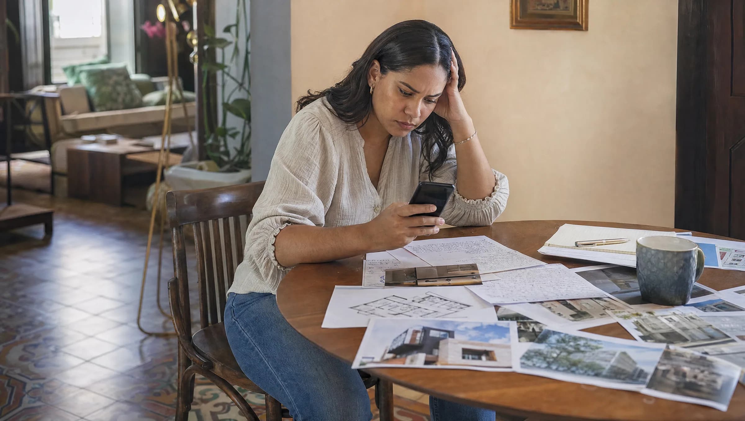 Mujer dominicana revisando con frustración anuncios de propiedades en su teléfono, rodeada de documentos y notas dispersas sobre su mesa