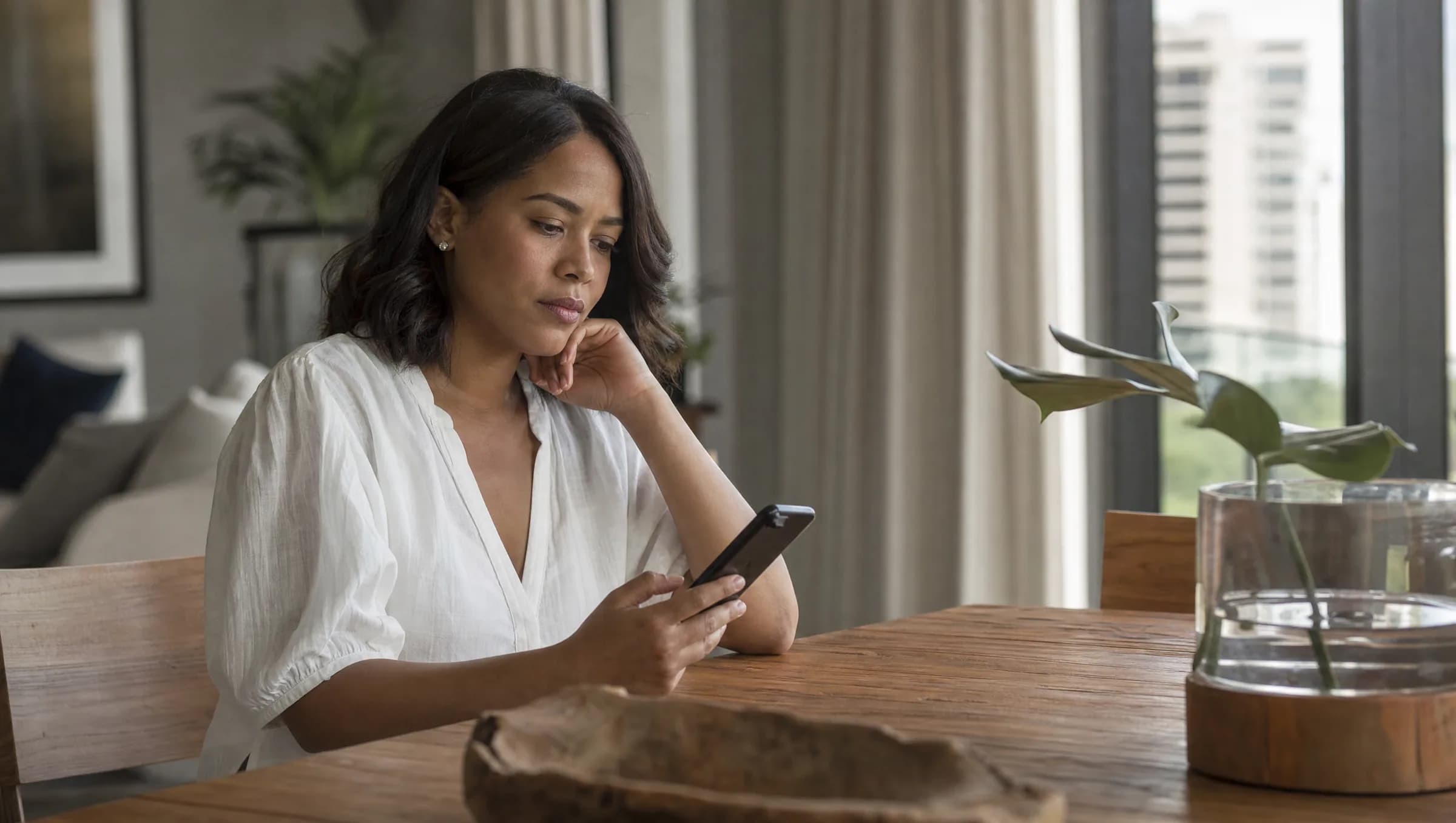 Mujer dominicana reflexionando mientras revisa ofertas inmobiliarias en su teléfono móvil en un apartamento en Piantini
