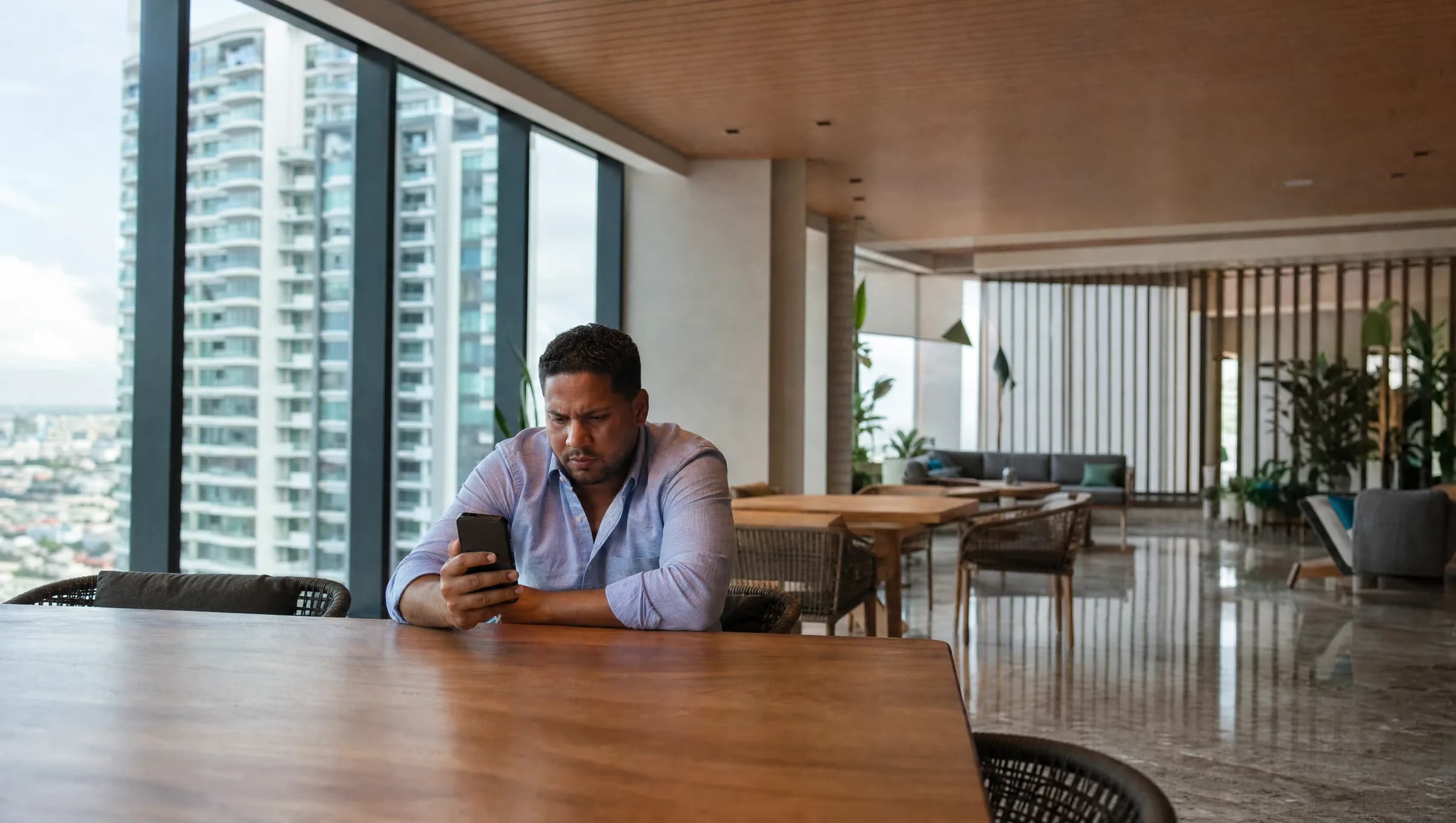 Hombre dominicano revisando preocupado los gastos de mantenimiento en su teléfono mientras está sentado en el área social vacía de su condominio en Santo Domingo