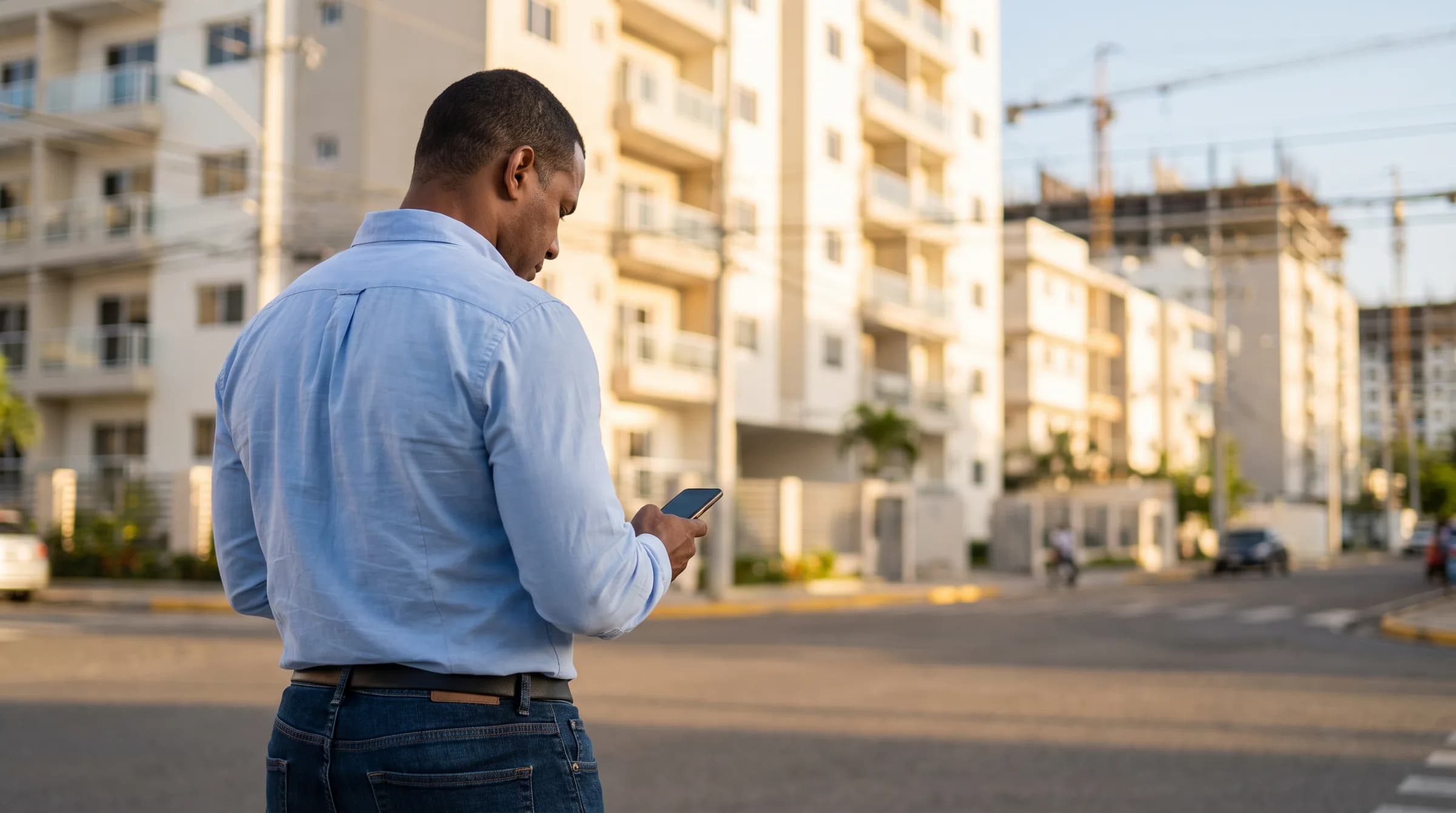 Dominicano contemplando decisiones inmobiliarias mientras revisa información en su teléfono frente a desarrollos urbanos en Santo Domingo