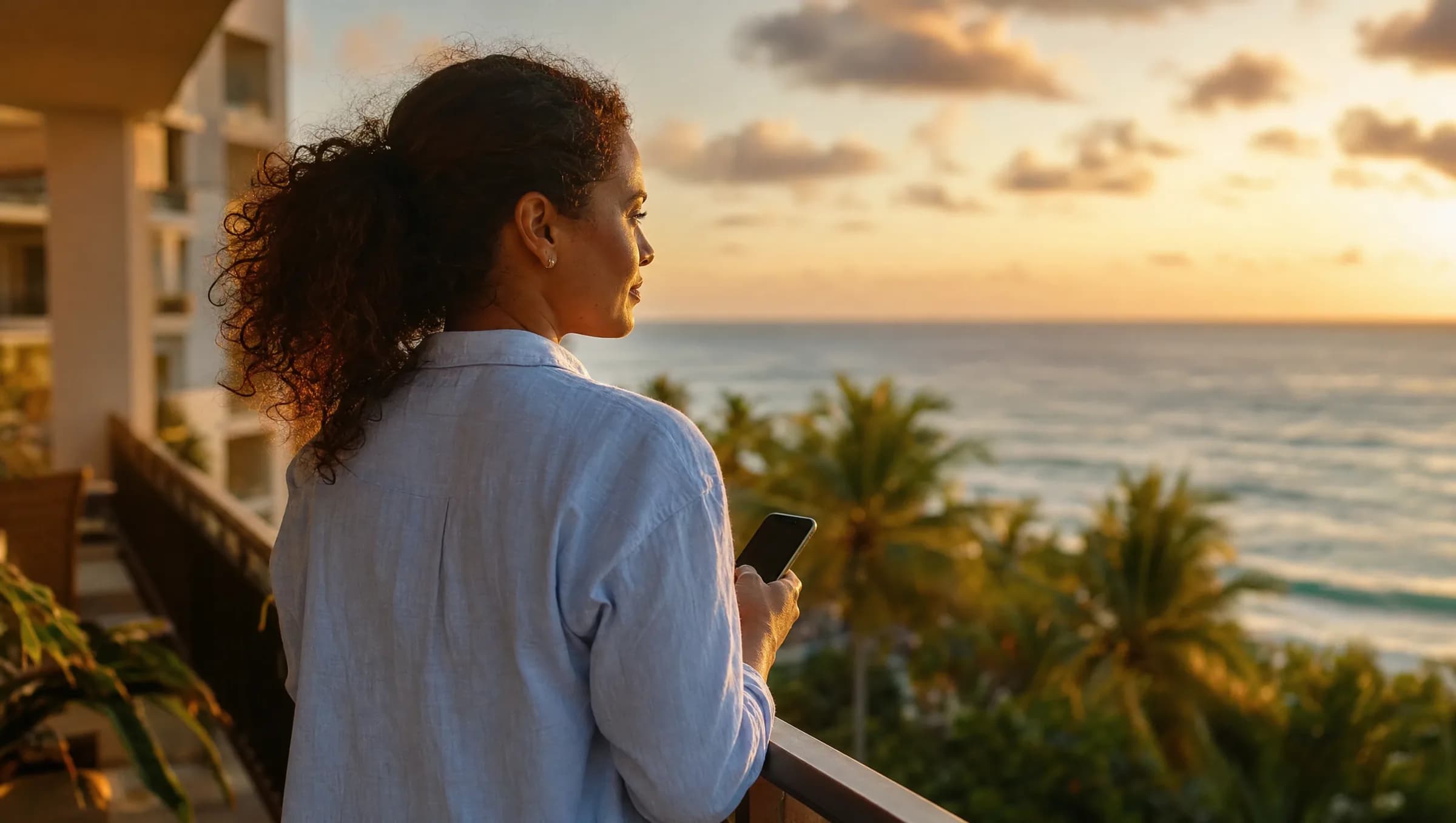 Mujer dominicana contemplando el atardecer desde un balcón en Punta Cana mientras revisa propiedades en su teléfono, reflexionando sobre inversiones inmobiliarias en zona turística