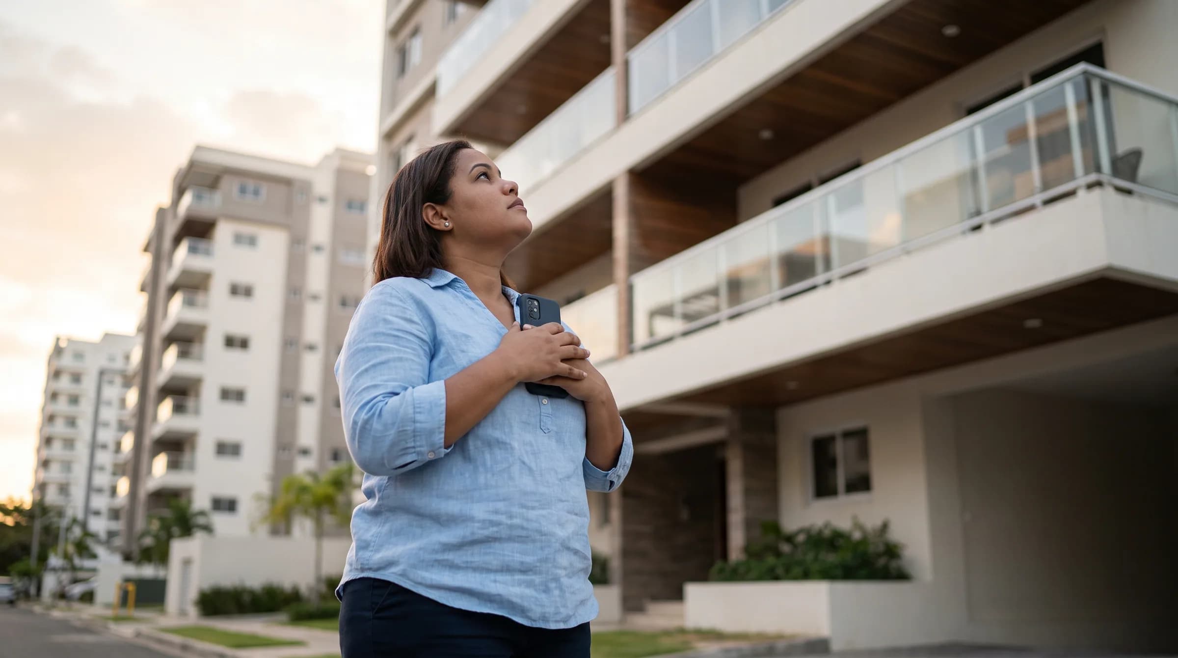 Mujer dominicana contemplando pensativamente un edificio residencial moderno en Naco mientras sostiene su teléfono, reflexionando sobre su decisión de compra