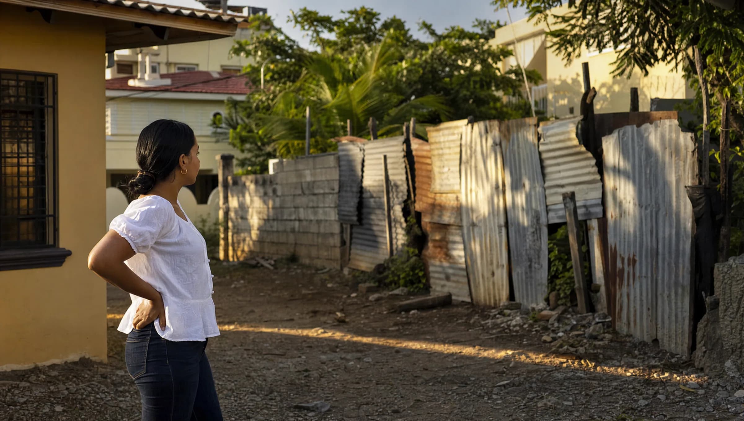 Mujer dominicana observando pensativamente su terreno en Santiago, con una verja al fondo que debe ser demolida debido a una servidumbre de paso no conocida al momento de la compra