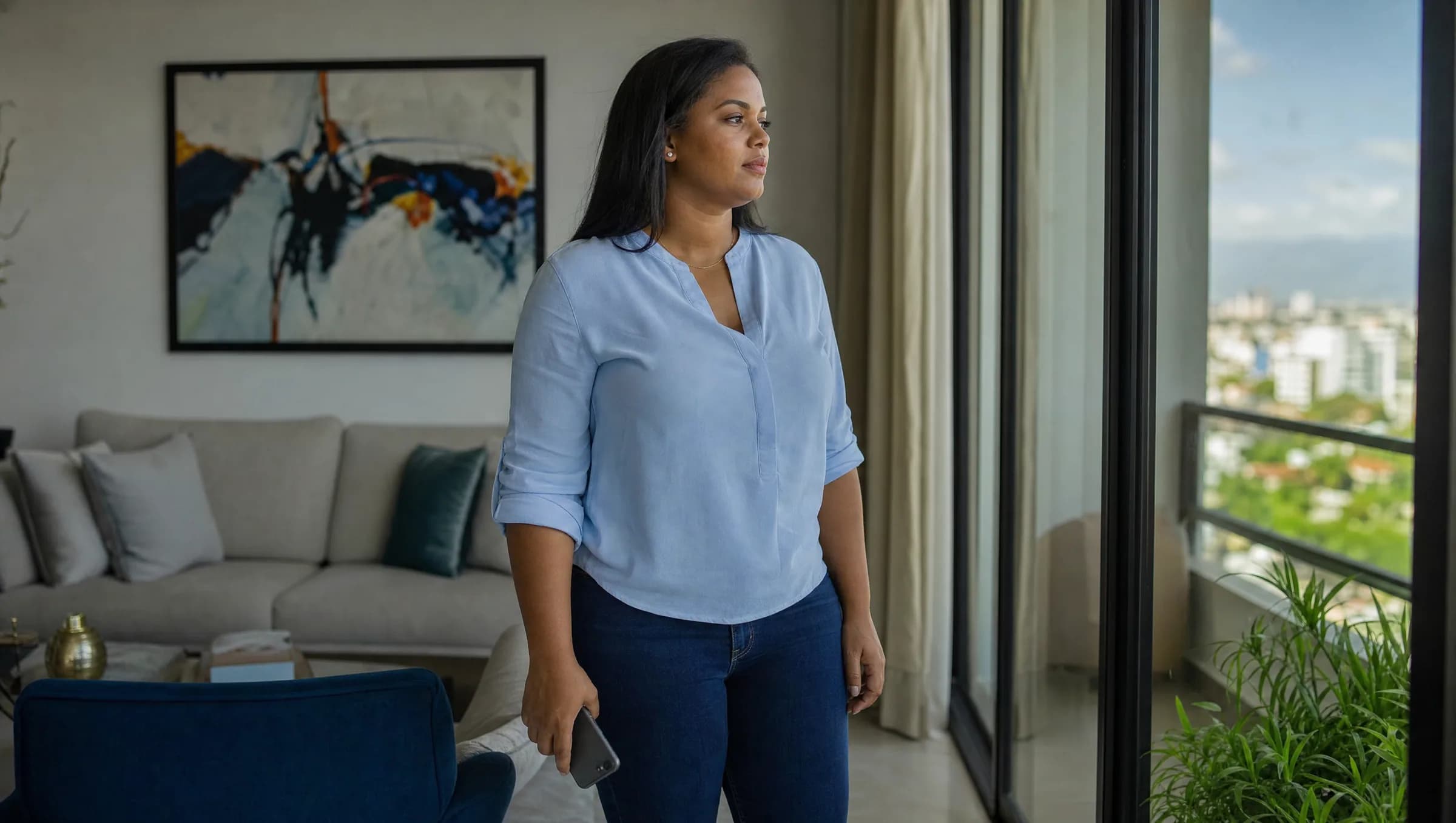 Mujer dominicana contemplativa en sala de apartamento moderno en Santiago, sosteniendo su teléfono mientras reflexiona sobre una decisión inmobiliaria importante